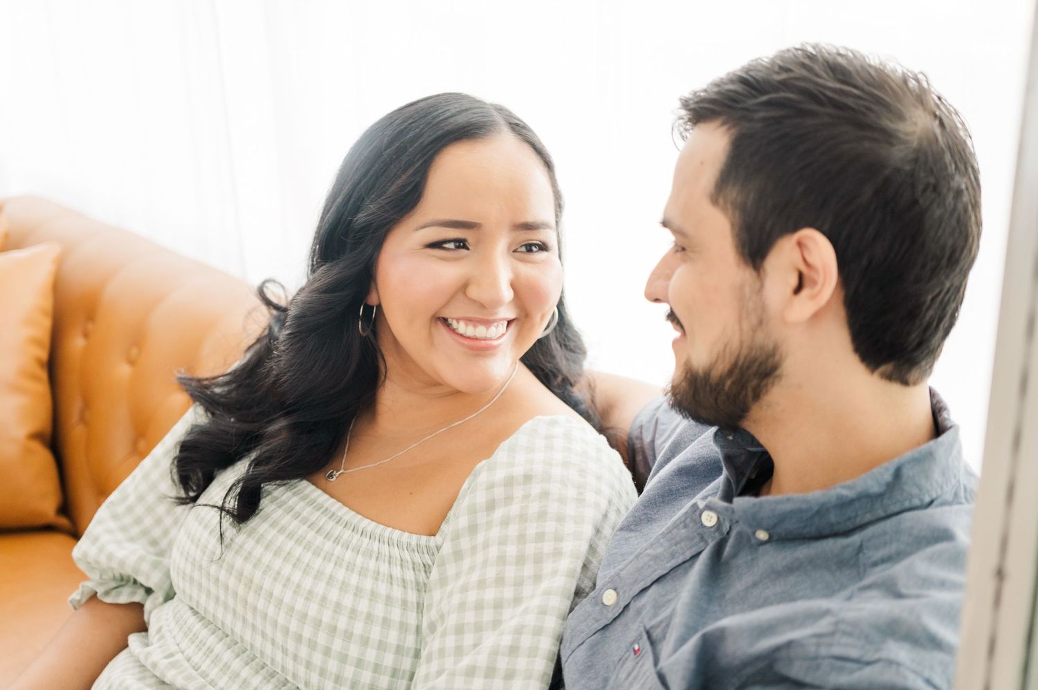 A couple shares a joyful moment while sitting on a tan leather couch in a bright room.