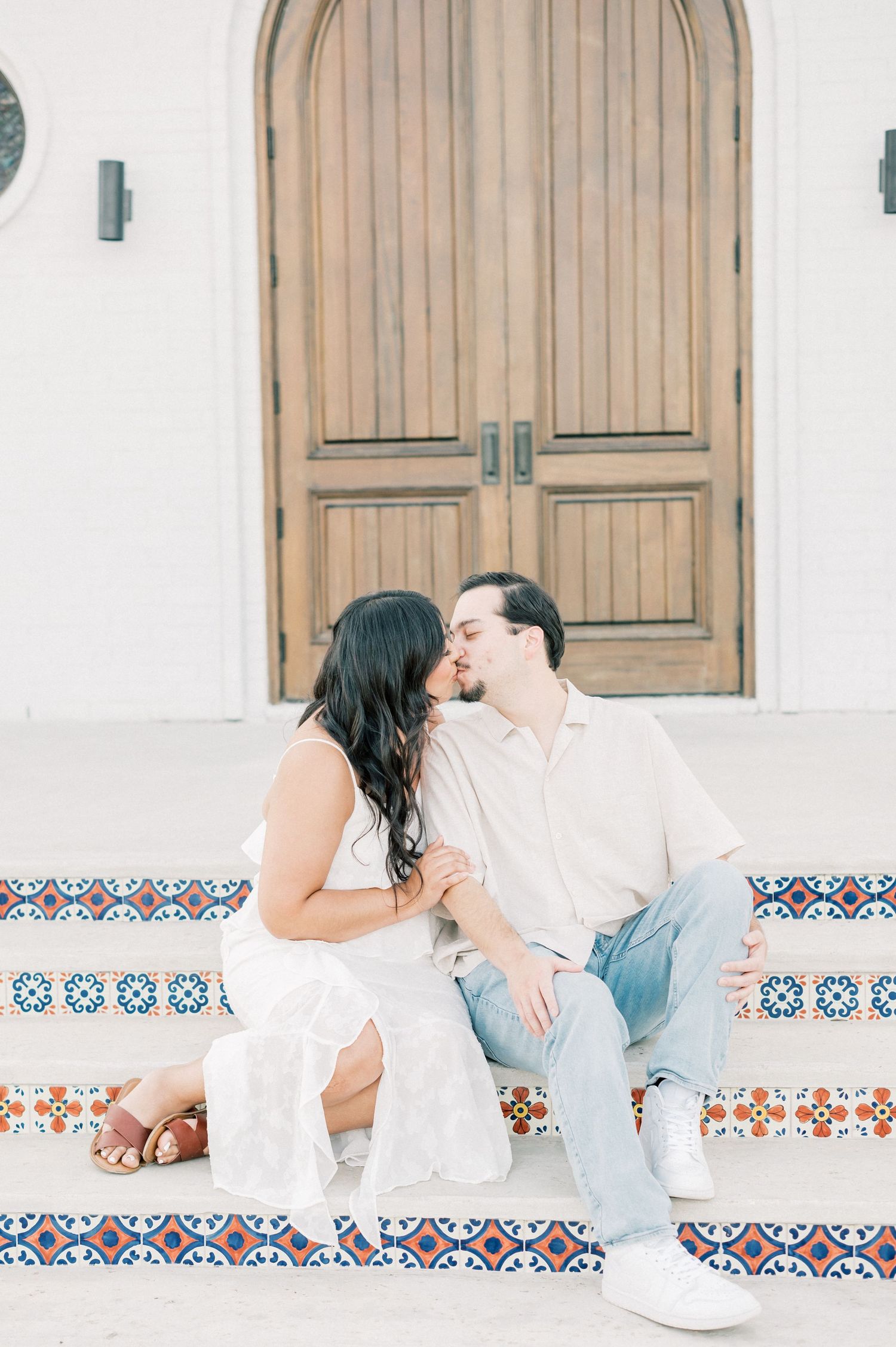 A couple in white and blue outfits share romantic moments on a decorative tiled bench in a Mediterranean setting.