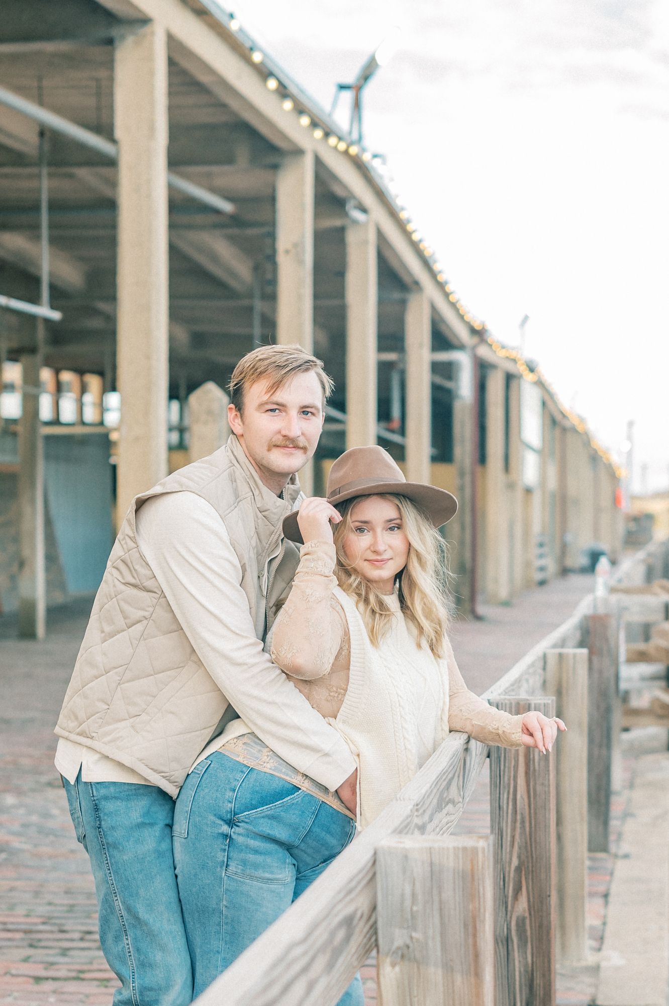 A couple in western-style clothing poses together near rustic wooden pillars and fencing.