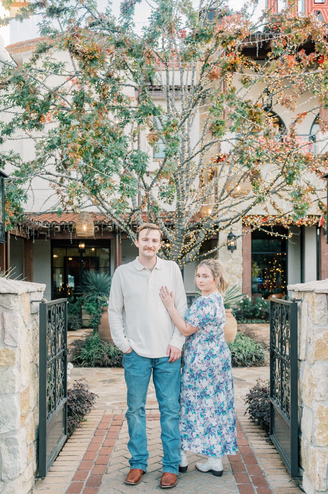 A couple poses together on a brick walkway decorated with twinkling lights and greenery.