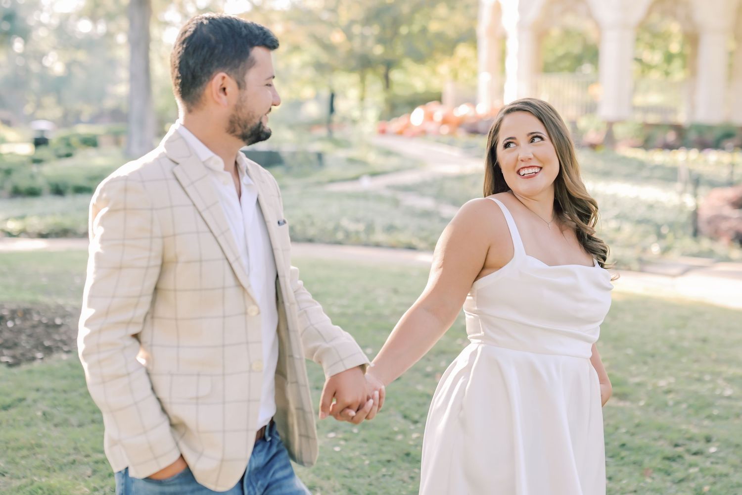 Couple in a sunny park holds hands while walking, the woman in a white dress smiling back at a man in a plaid blazer.