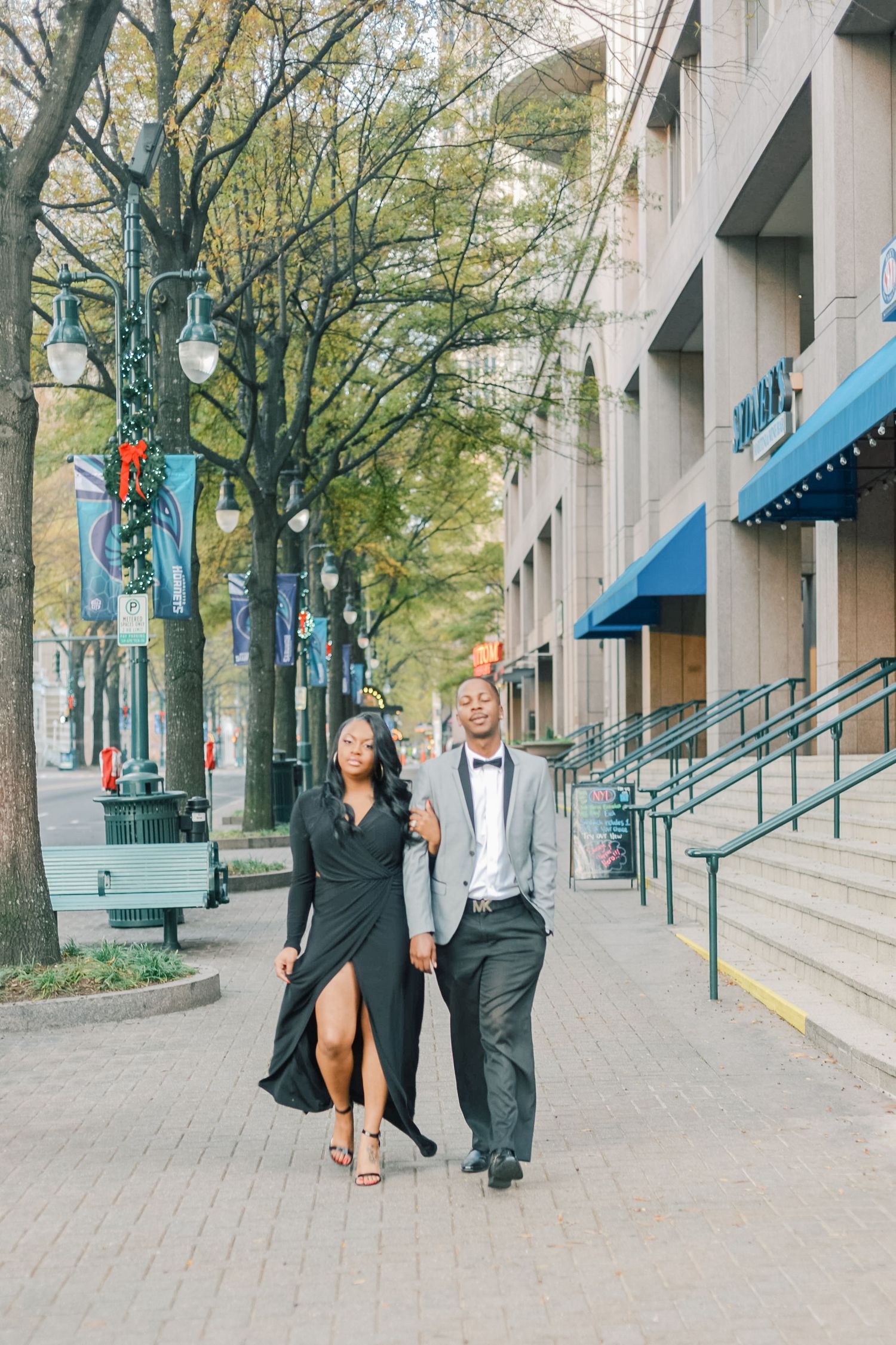 Elegant couple walking hand-in-hand on a city sidewalk, woman in black gown and man in gray suit with bow tie.