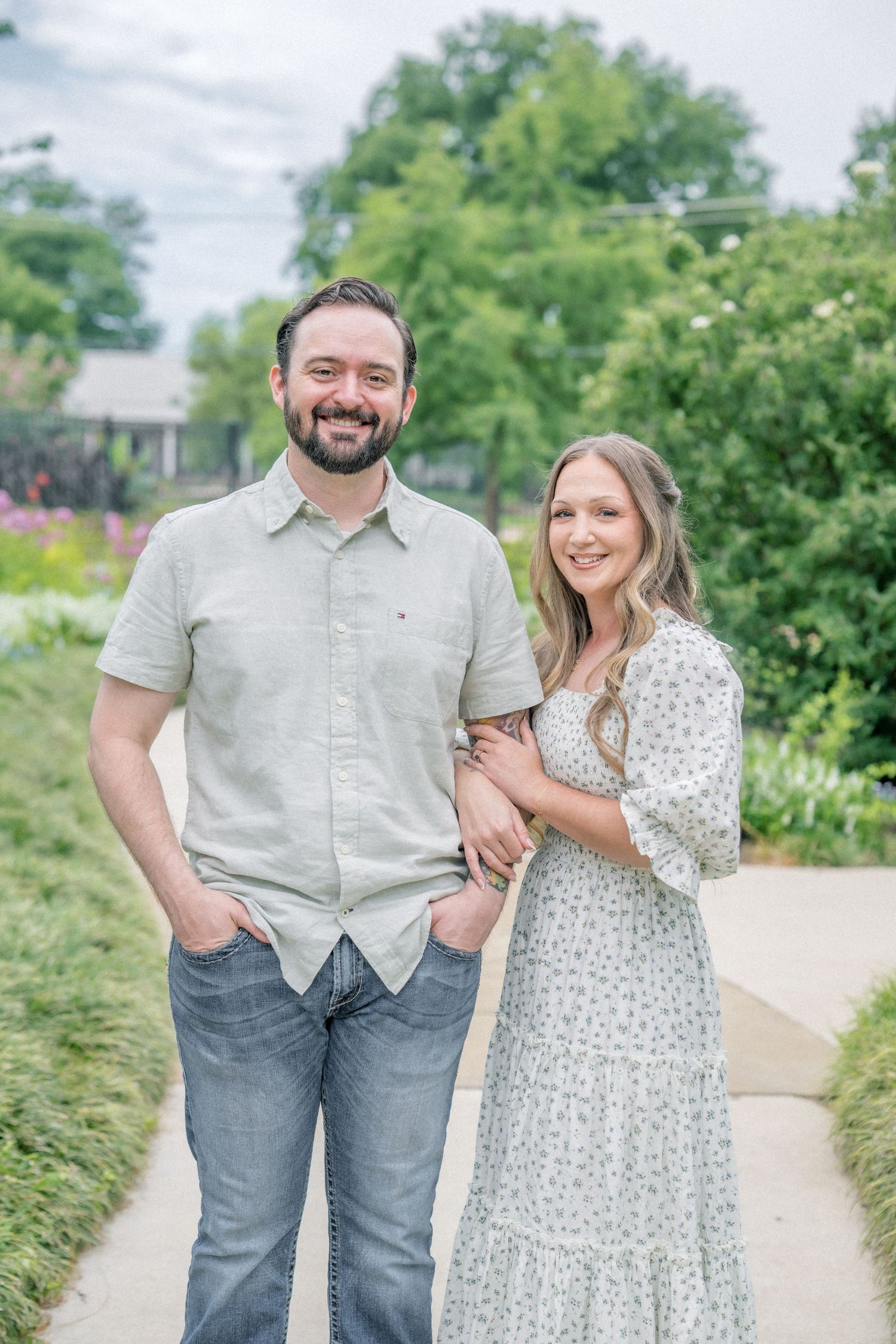Smiling couple poses together on a garden path, man in gray shirt and jeans, woman in floral maxi dress.