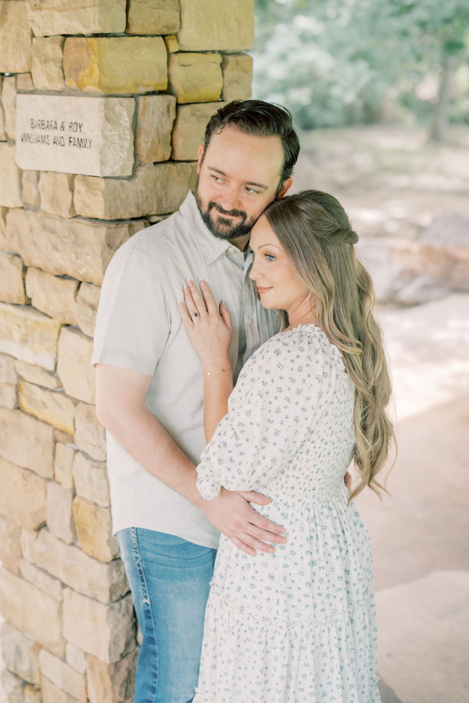Couple posing tenderly against a rustic stone pillar in a sunlit garden setting.