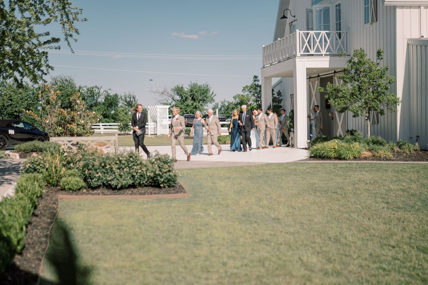 Wedding guests mingling outside a white barn venue surrounded by manicured landscaping on a sunny day.