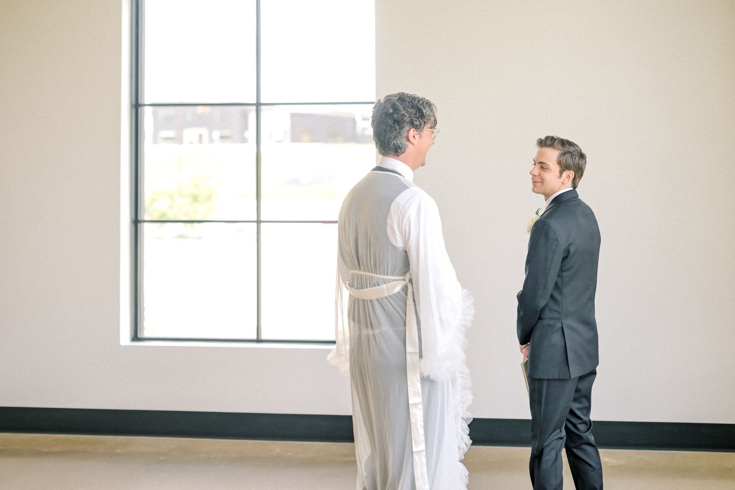 Bride in white gown sharing quiet moment with officiant in white robe near large window before ceremony.