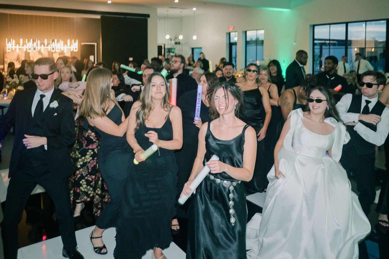 Lively wedding reception dance floor filled with guests in black formal attire and bride in white gown.