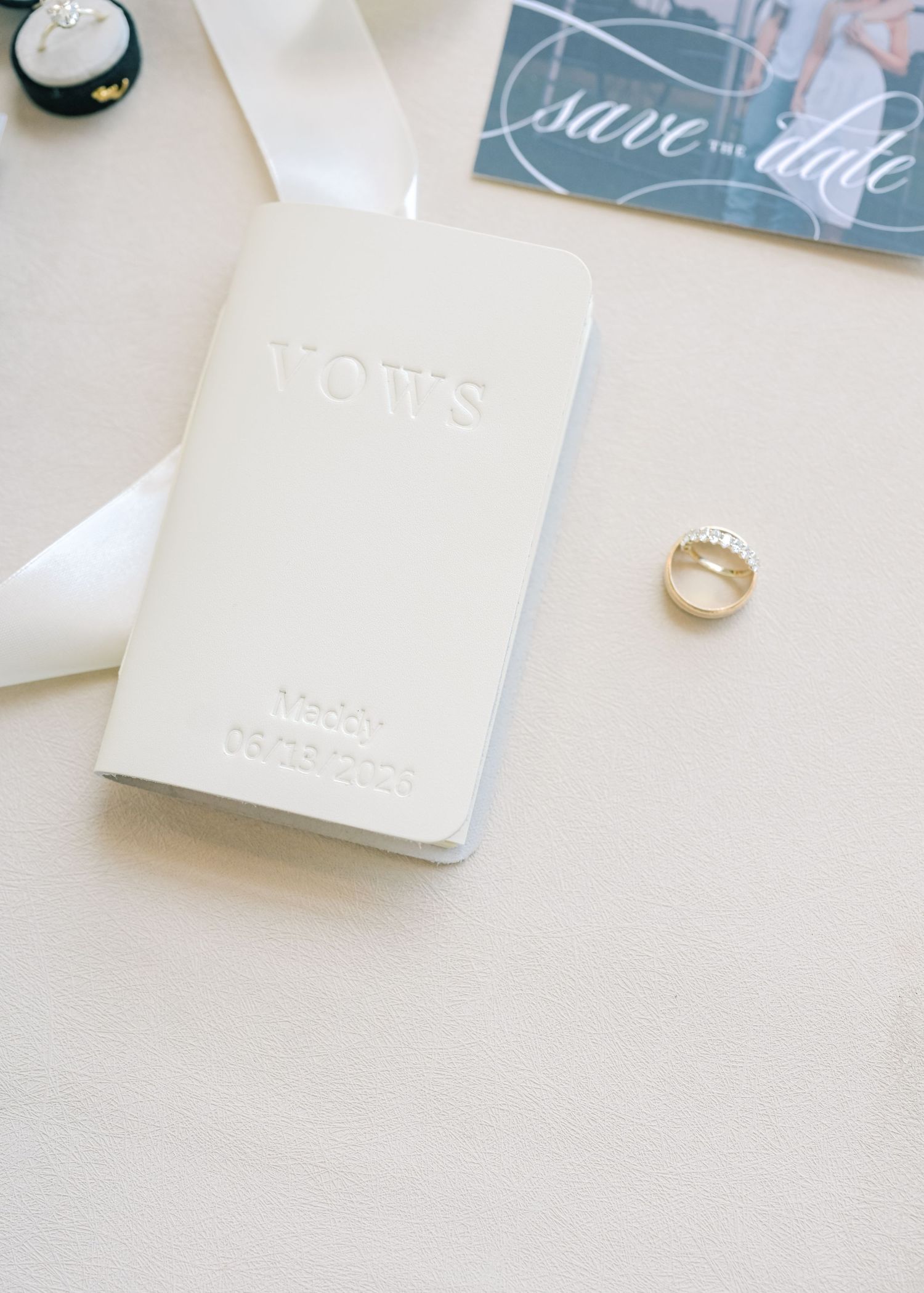 White leather vow book personalized with name and date displayed with gold wedding band and save the date.