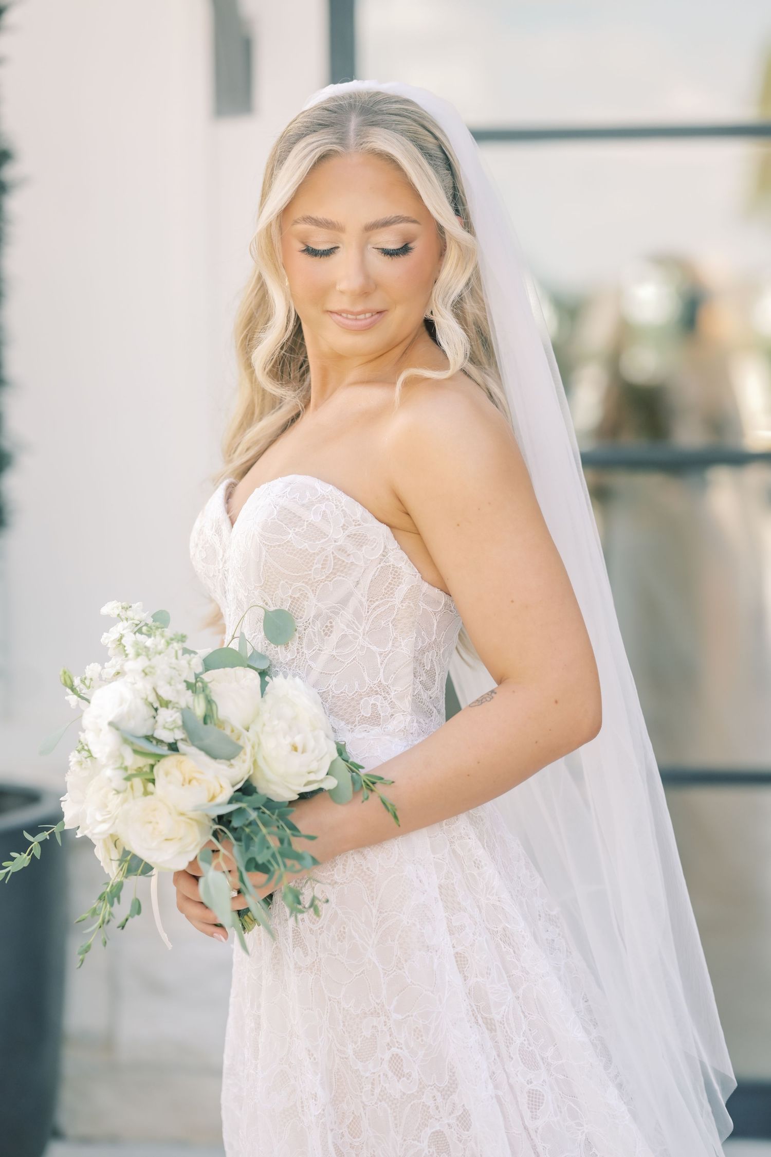 Elegant bride in strapless lace gown and veil holds white rose and eucalyptus bouquet.