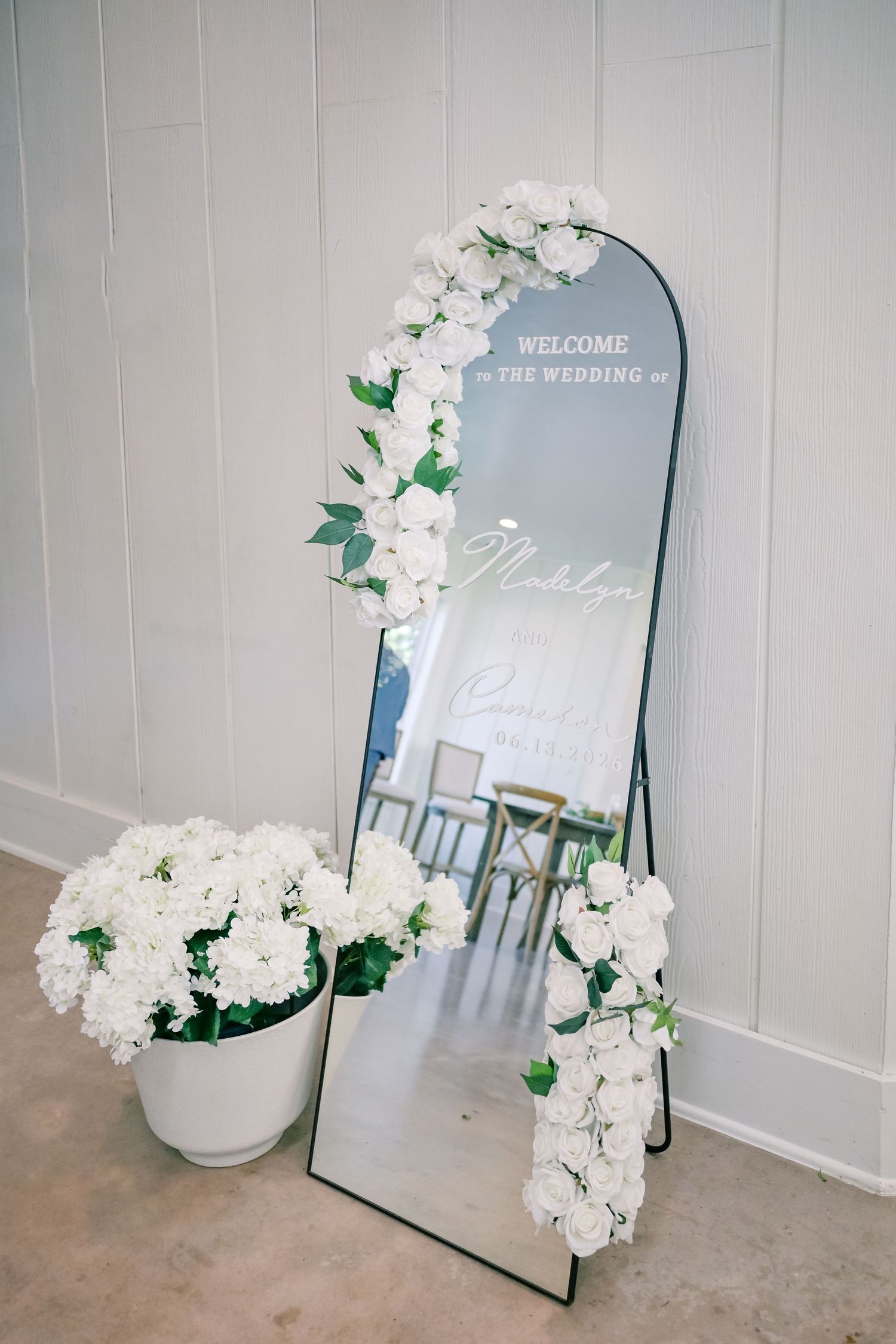 Elegant wedding welcome mirror decorated with white floral garland beside white hydrangea arrangement.