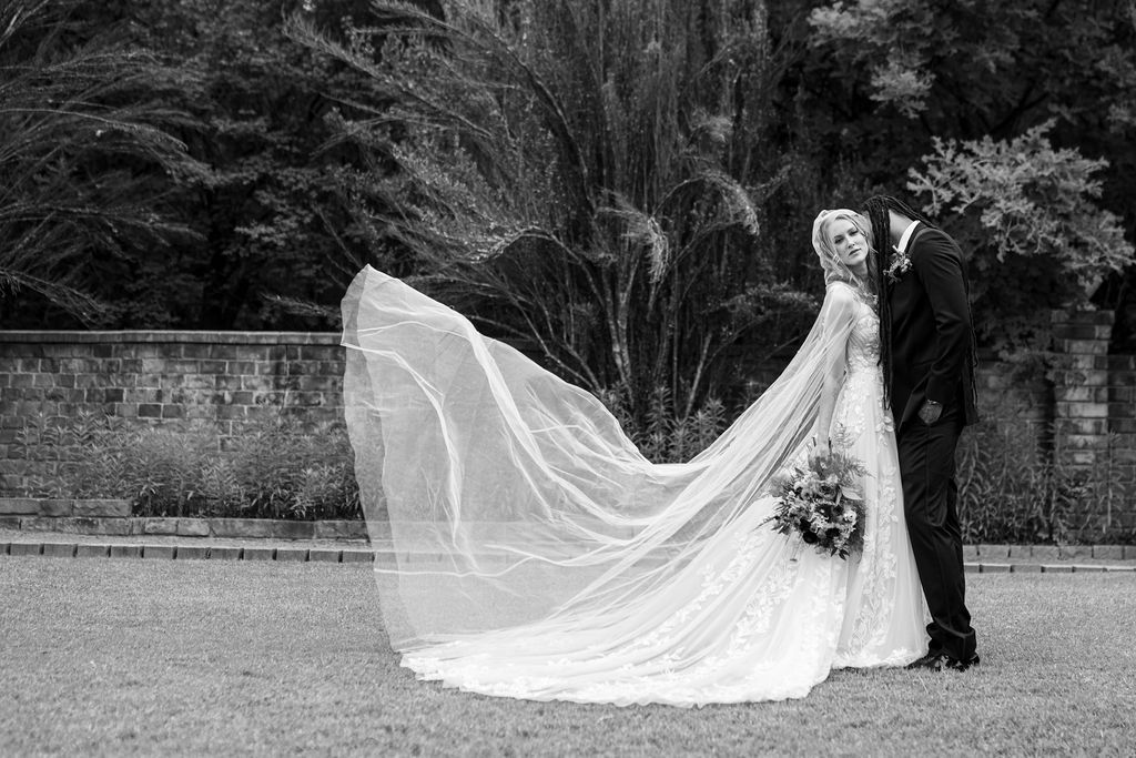 Black and white wedding photo of bride in flowing veil and groom in tuxedo kissing outdoors near brick wall.