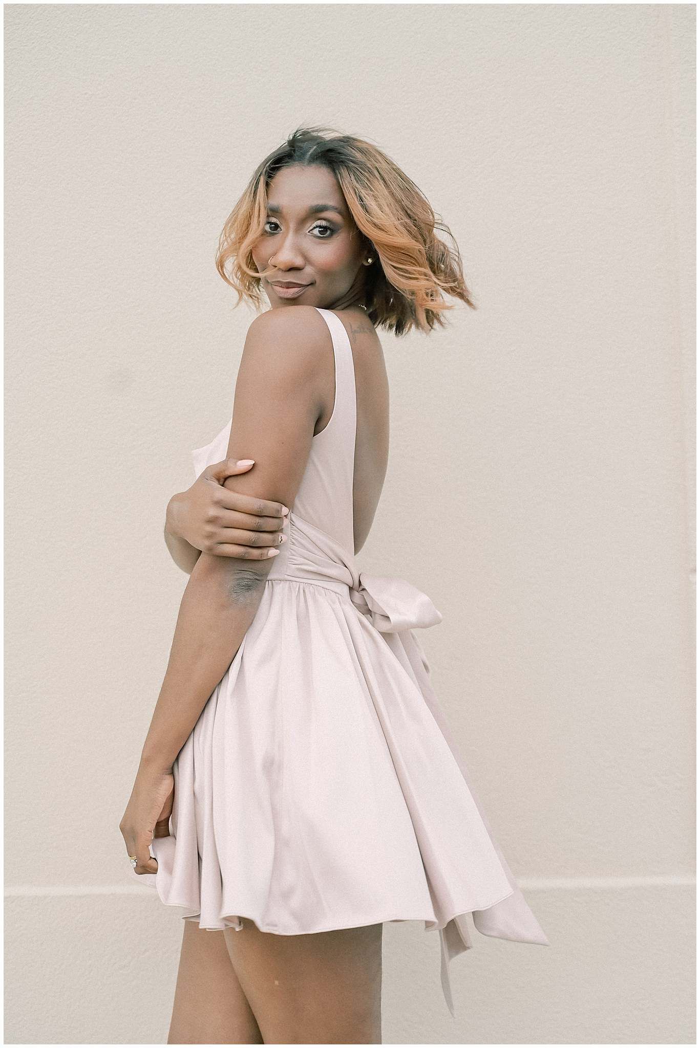 A woman in a flowy pink dress with a bow detail poses against a neutral wall while looking over her shoulder.