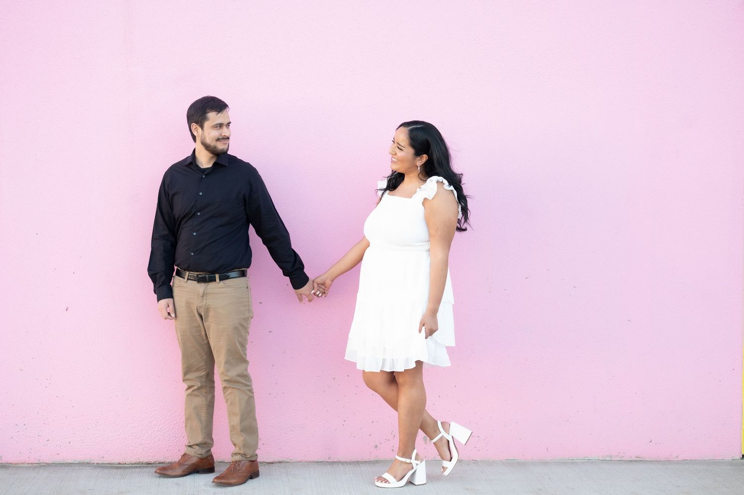 A romantic couple shares a playful moment against a pink wall while dressed in contrasting outfits.