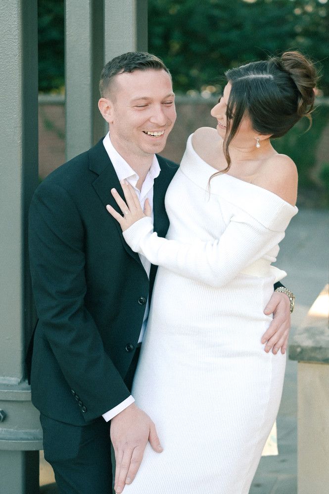 Smiling groom in dark suit embraced by bride in off-shoulder white dress during outdoor wedding portrait.