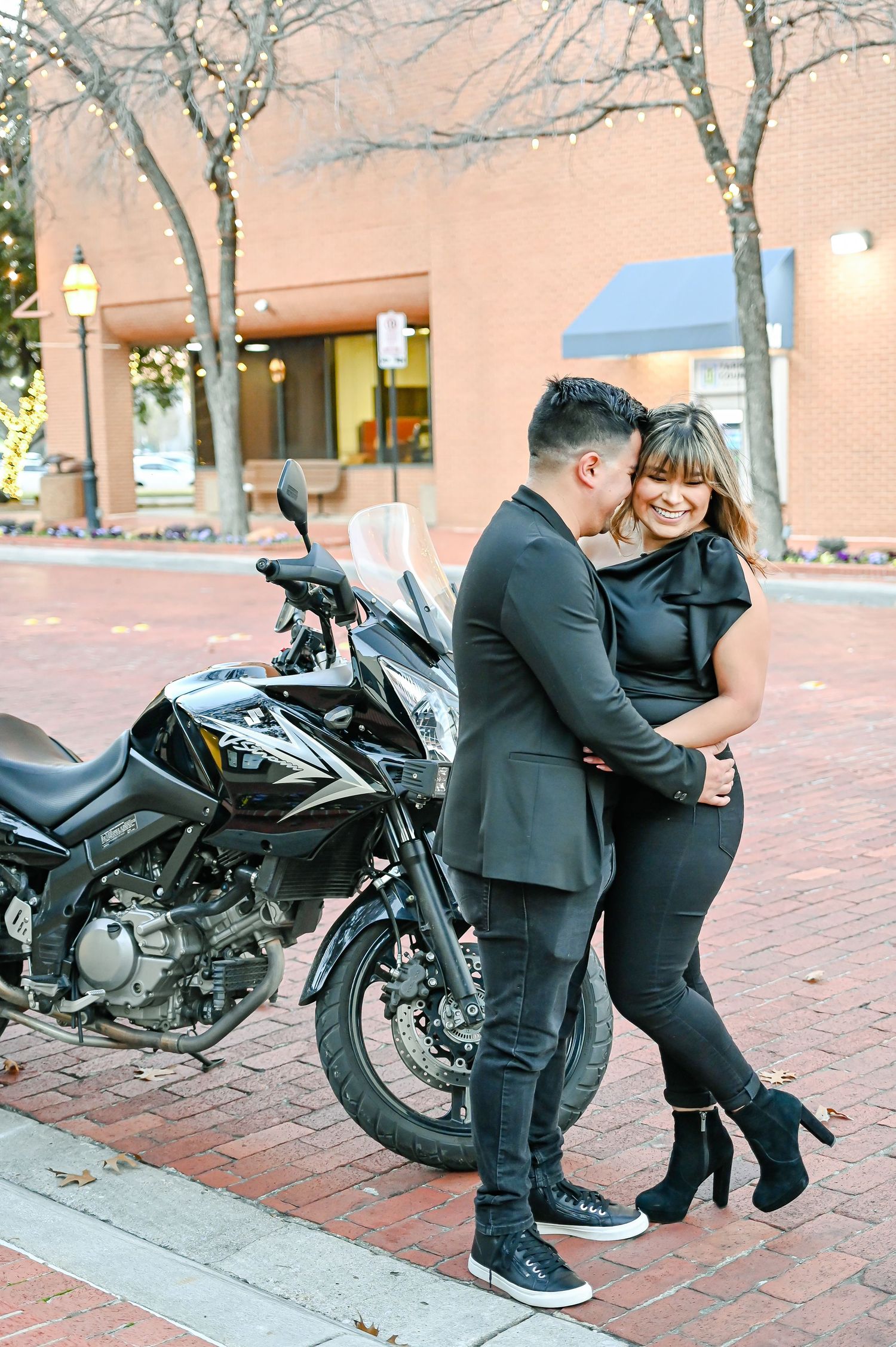 Romantic couple in black attire laughing together beside a parked motorcycle outdoors.