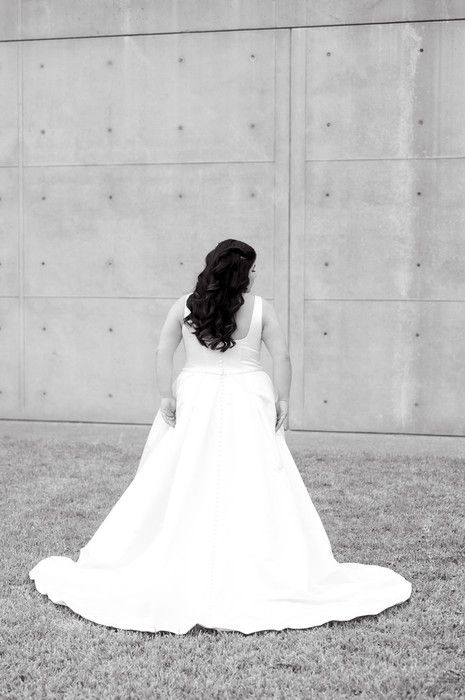 Bride in elegant white gown with long curly hair facing concrete wall.
