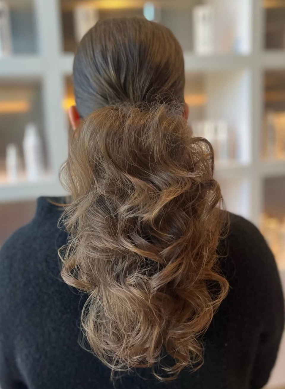 Woman with elegant voluminous curly ponytail hairstyle, viewed from behind in a salon setting.