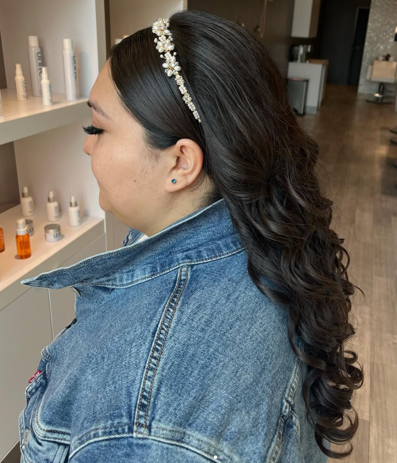 Woman with dark curly ponytail wearing a pearl headband and denim jacket in a hair salon.