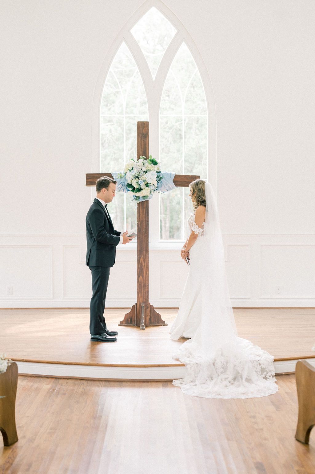 Bride and groom exchange vows at altar with wooden cross and floral arrangement in bright white chapel.