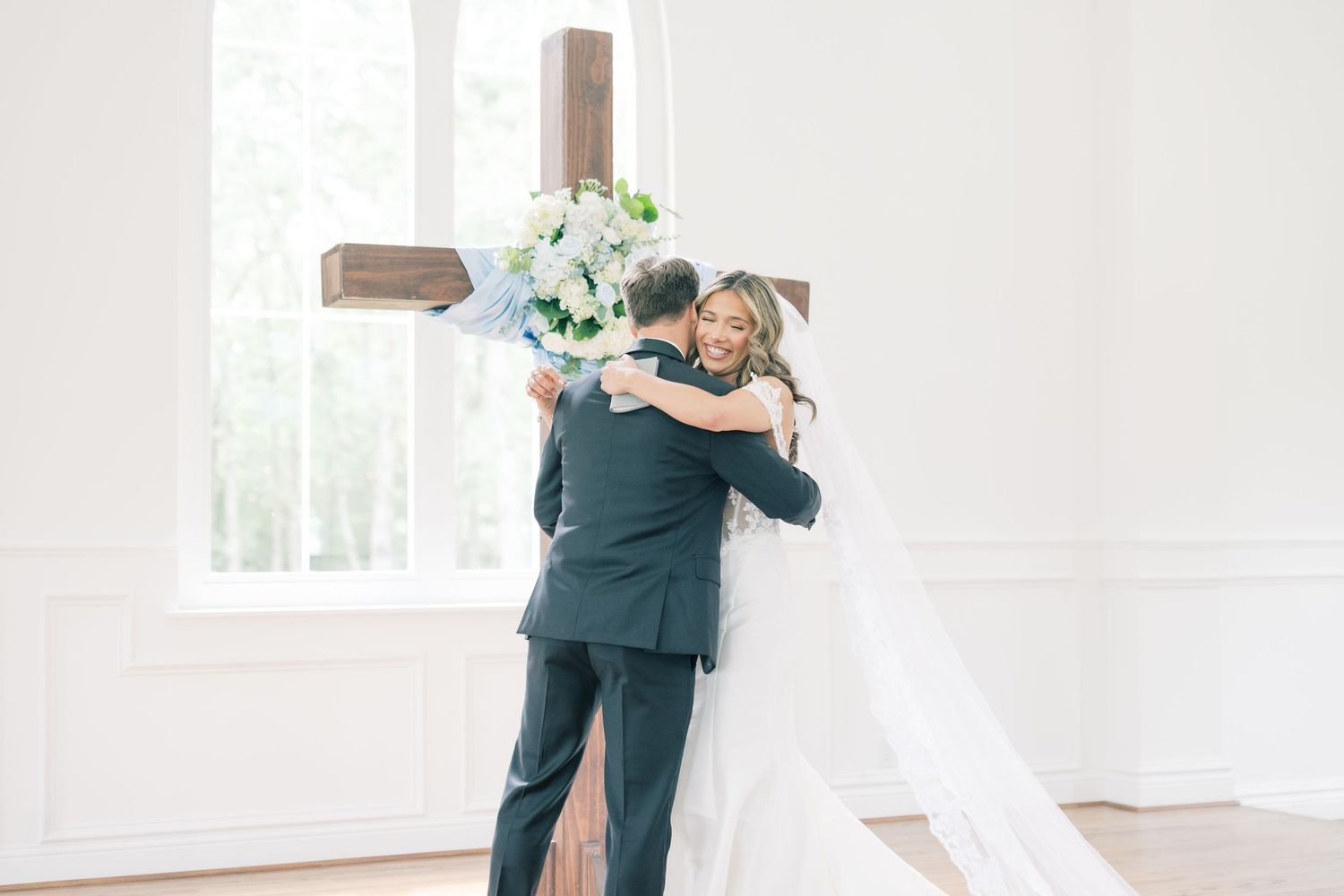 Bride and groom embrace in front of a wooden cross in a sunlit white wedding chapel.