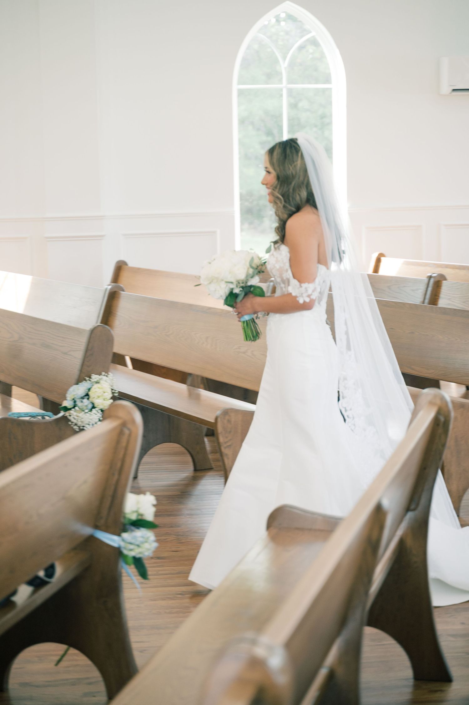 Bride in a white off-shoulder gown with long veil holding a white bouquet inside a bright white chapel.