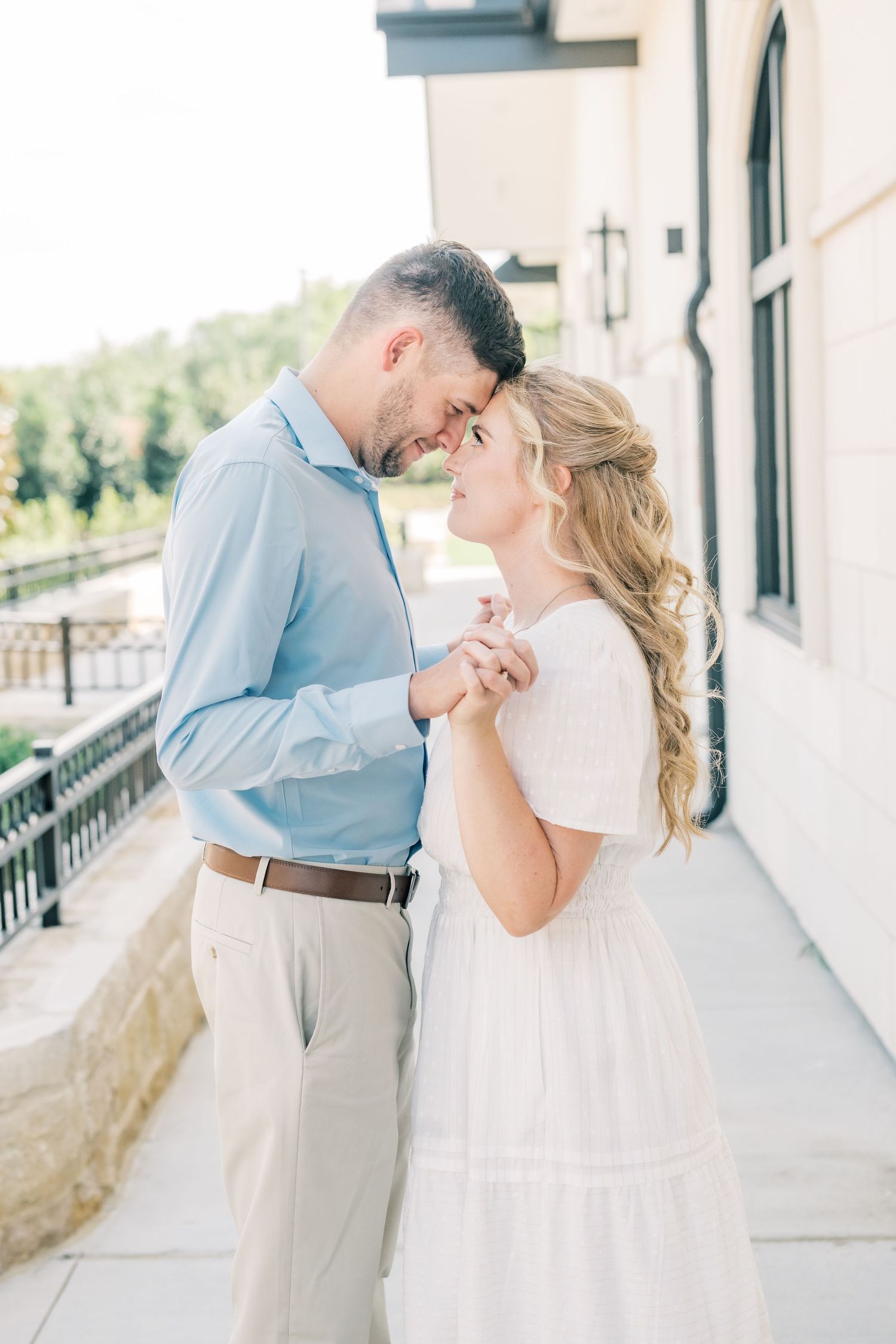 A romantic couple holds hands and touches foreheads tenderly on a bright outdoor balcony.
