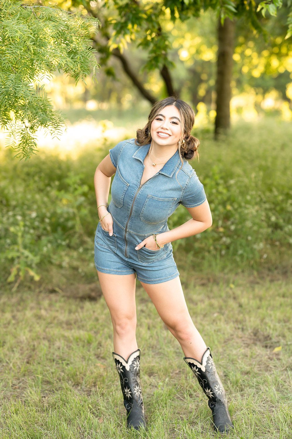 Smiling young woman in denim romper and cowboy boots posing outdoors in a lush green park setting.