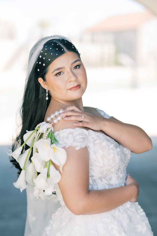 A bride wearing a beaded headpiece and off-shoulder wedding dress poses with white calla lily bouquet.