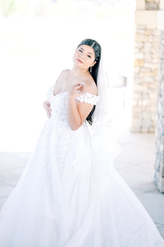 A bride in a flowing white ball gown poses gracefully against a light stone wall in an elegant outdoor setting.