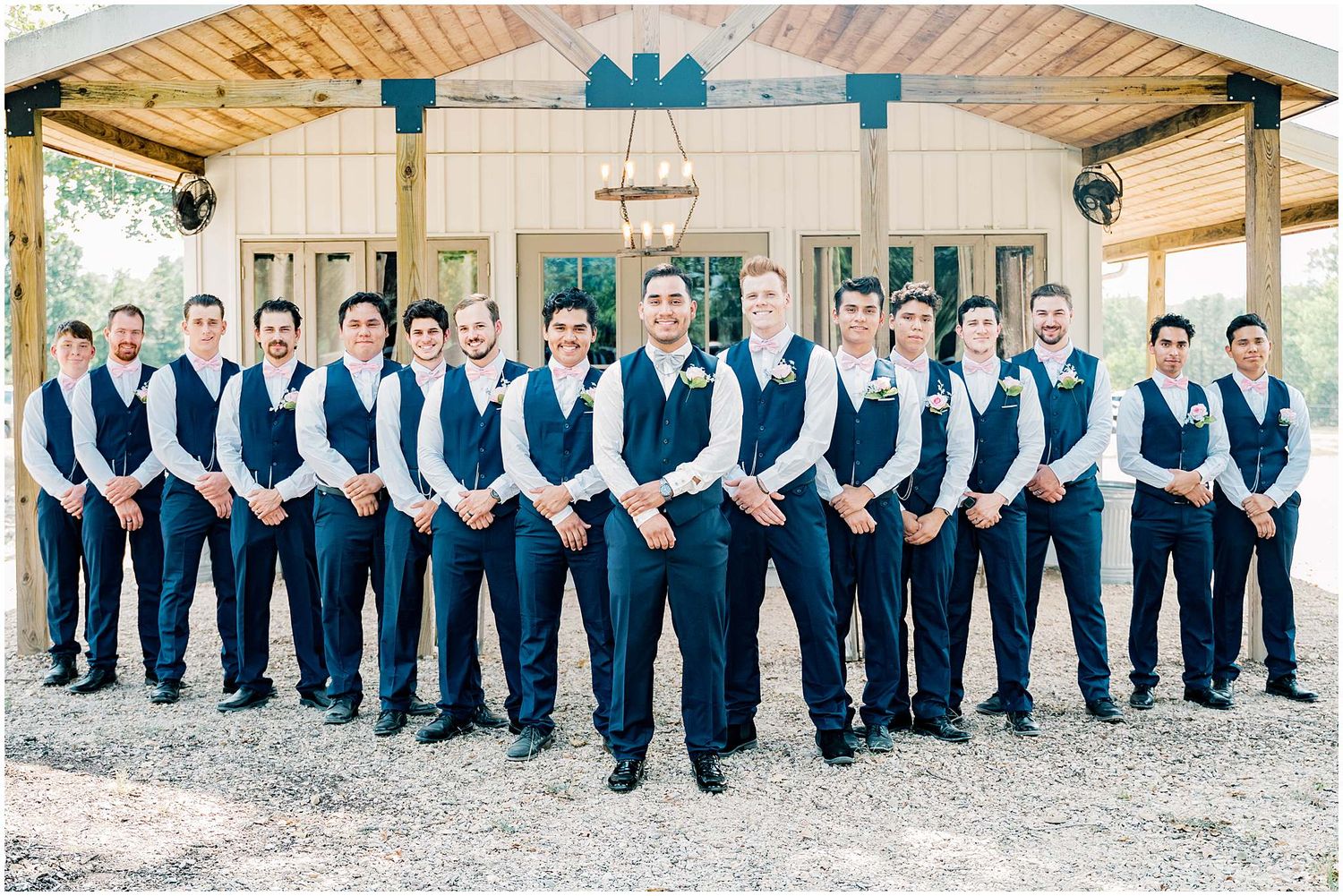 A line of groomsmen wearing matching navy blue vests and pants stand in formation under a rustic wooden pergola.