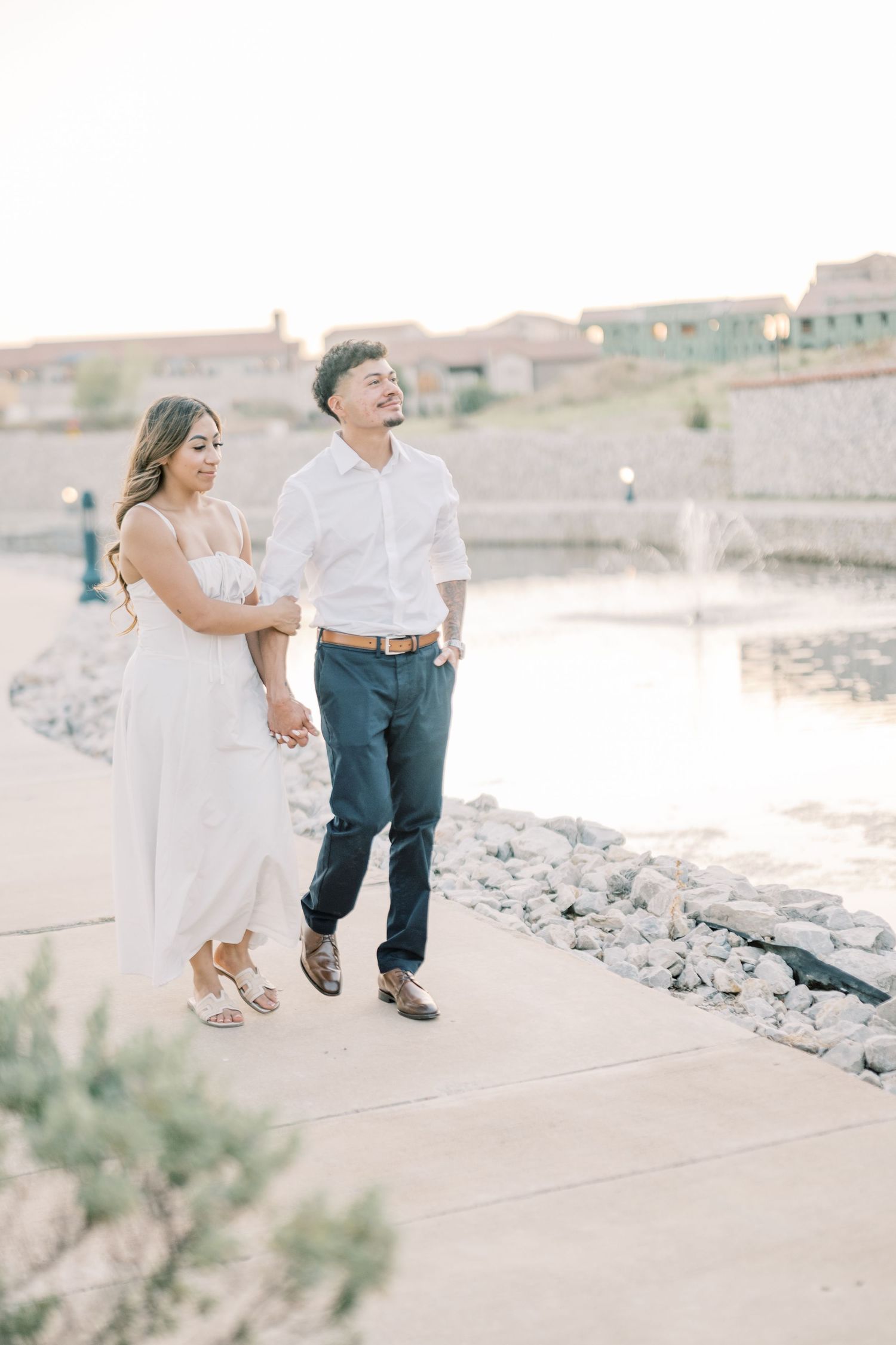 A happy couple walks hand in hand along a waterfront path during their golden hour engagement session.