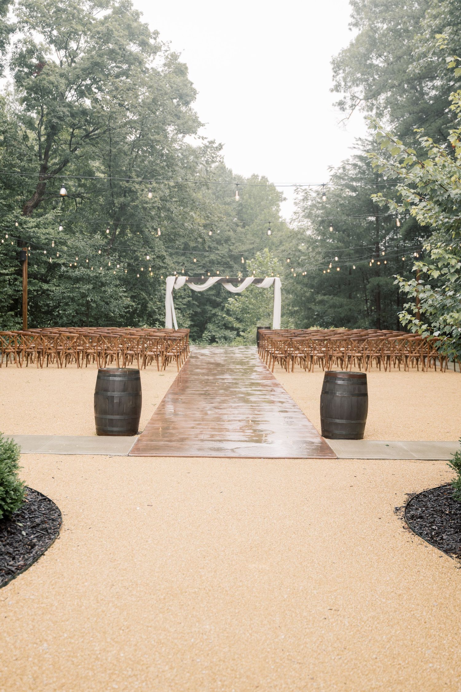 Outdoor wedding ceremony aisle with wooden barrel decor, string lights, and white floral arch amid lush trees.