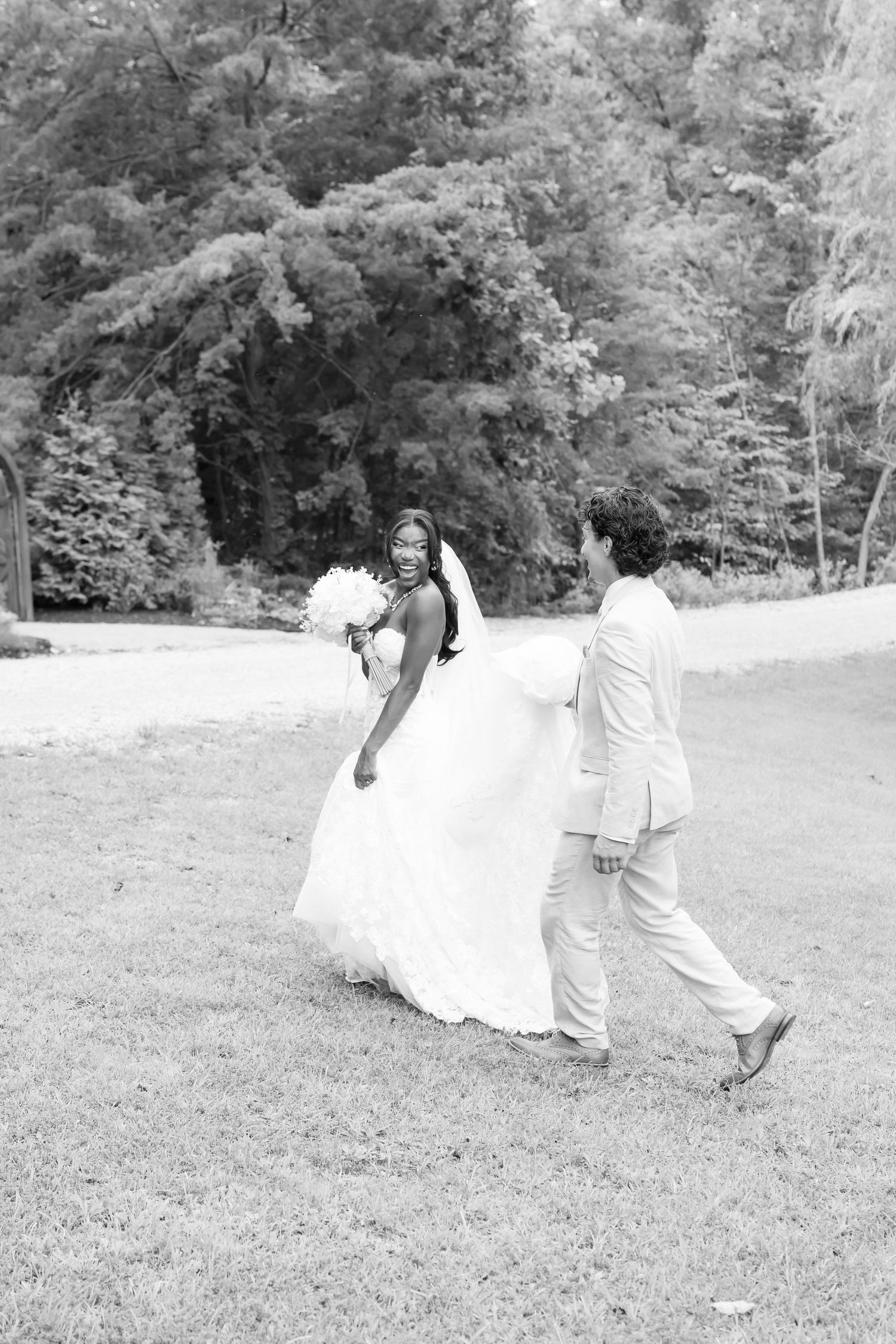 Black and white photo of a bride and groom walking on grass, bride holding bouquet, trees in background.