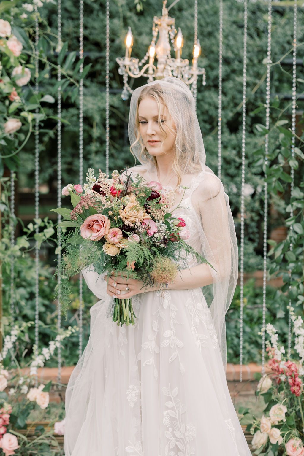 Elegant bride in lace gown holding colorful floral bouquet, standing beneath crystal chandelier surrounded by greenery.
