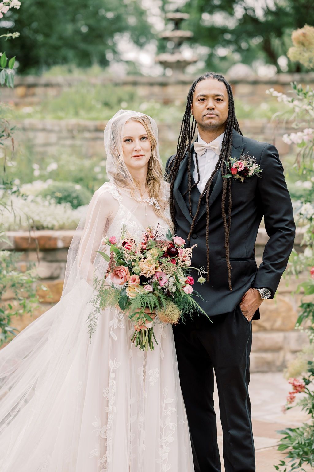 Bride in lace gown with veil holding floral bouquet stands beside groom in black suit with dreadlocks in garden.