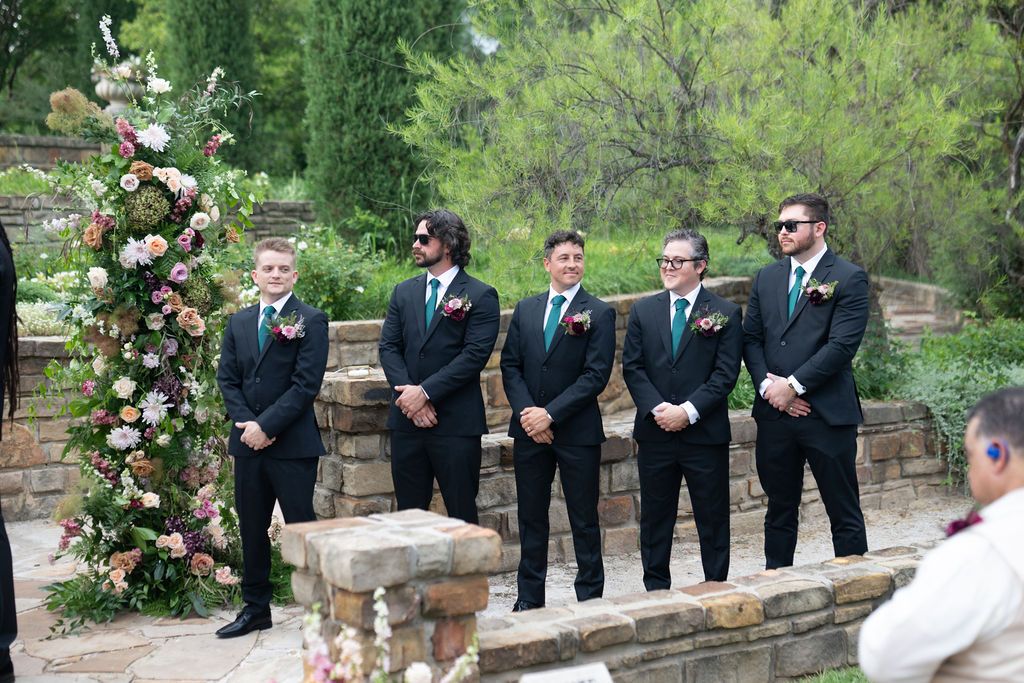 Five groomsmen in black suits with green ties stand at the altar during an outdoor wedding ceremony.