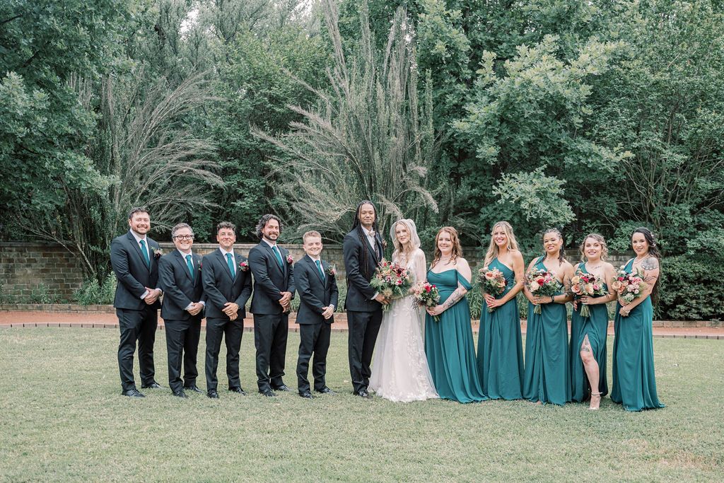 Wedding party posing outdoors with groomsmen in navy suits and bridesmaids in teal gowns, surrounded by lush green trees.