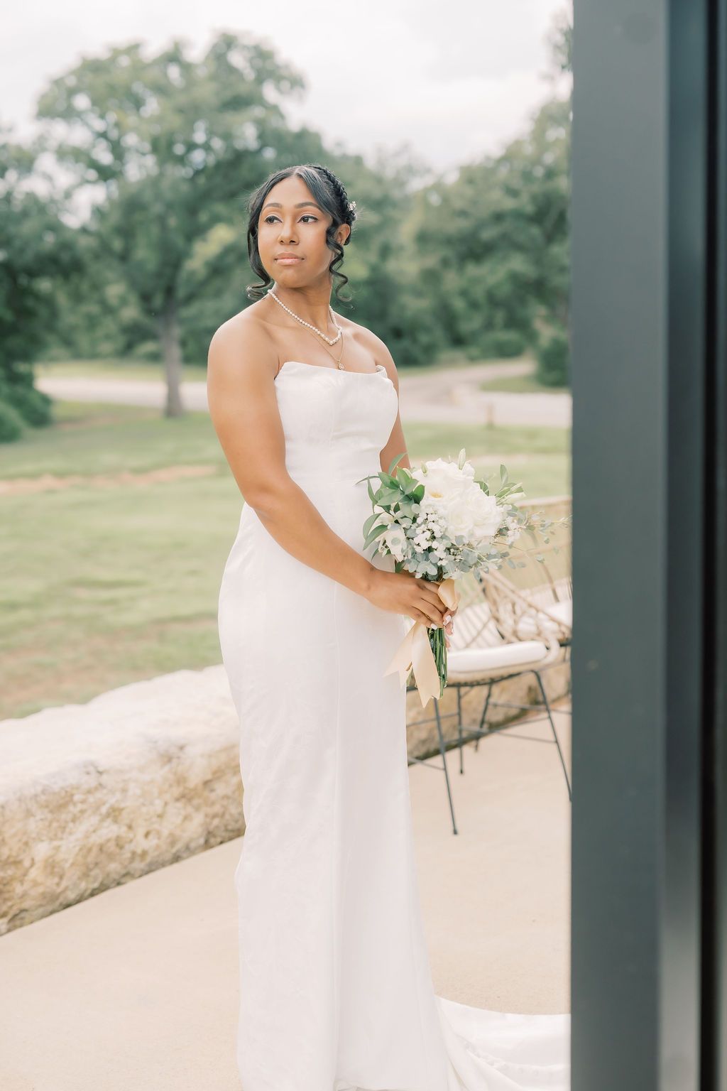 Elegant bride in a white strapless gown holding a white bouquet with greenery, standing outdoors near lush trees.