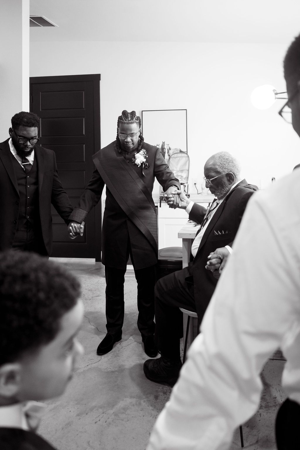Black and white photo of groom praying with groomsmen and elder before wedding ceremony.