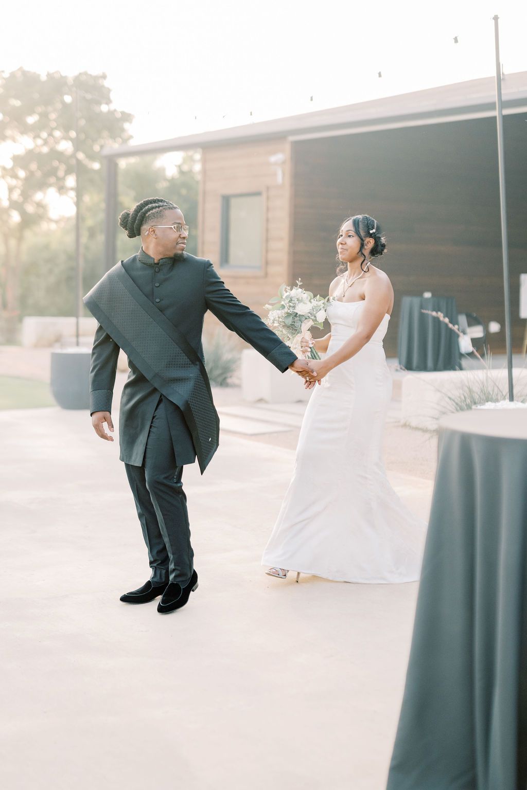 Groom in dark suit holds bride's hand as she carries white bouquet at outdoor wedding.