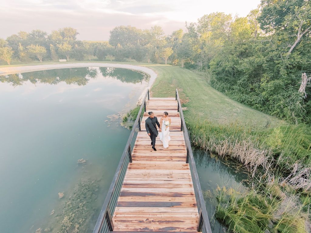 Aerial view of a wedding couple walking hand-in-hand on a wooden bridge over a serene pond surrounded by lush greenery.