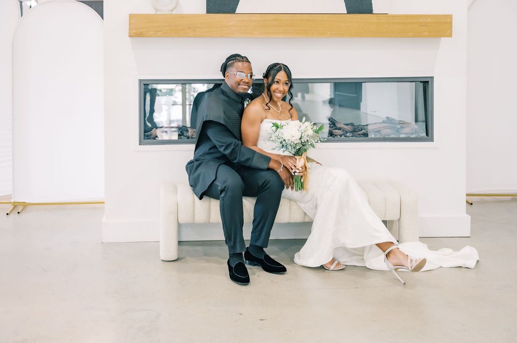 Bride and groom sit together on a white couch, smiling, bride holding a white bouquet.