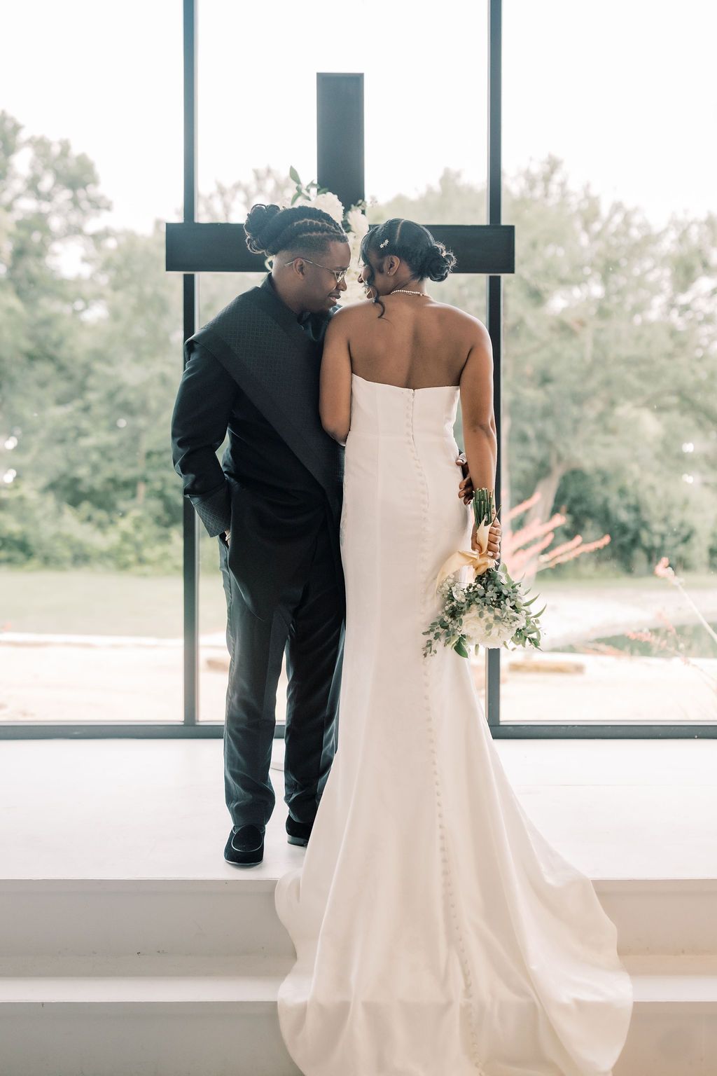 Bride and groom standing before a cross window, sharing an intimate moment in wedding attire with bridal bouquet.