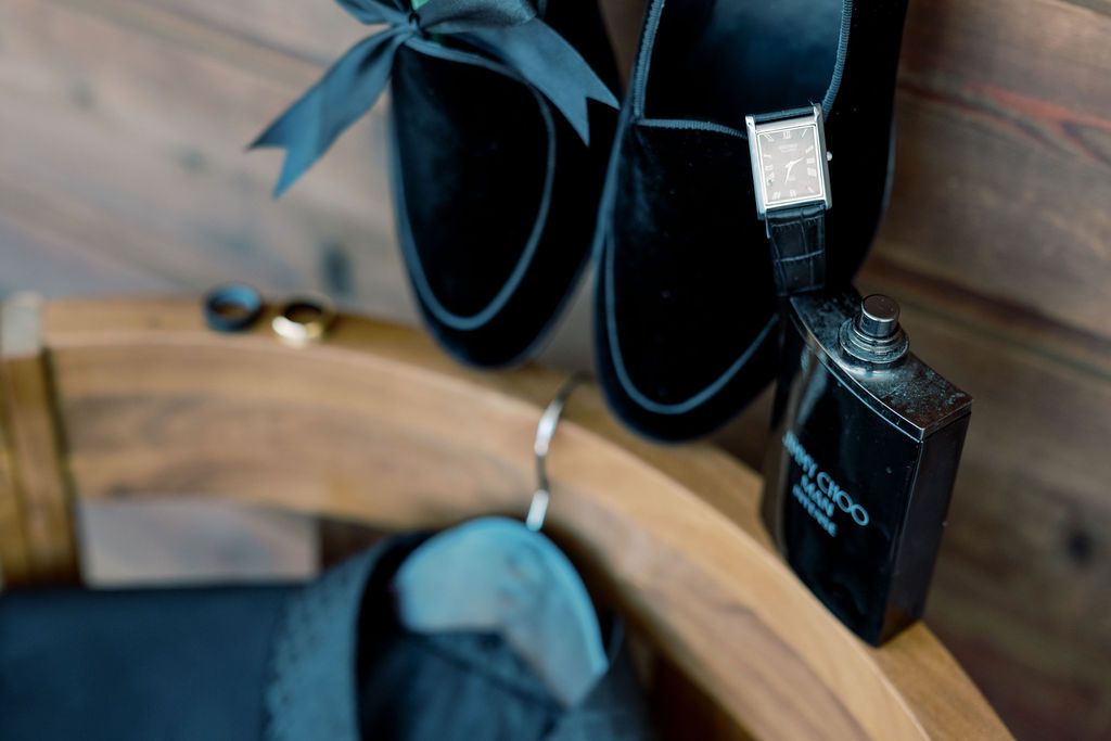 Black velvet dress shoes with bow tie, watch, and cologne arranged on wooden surface for a groom's wedding day flat lay.