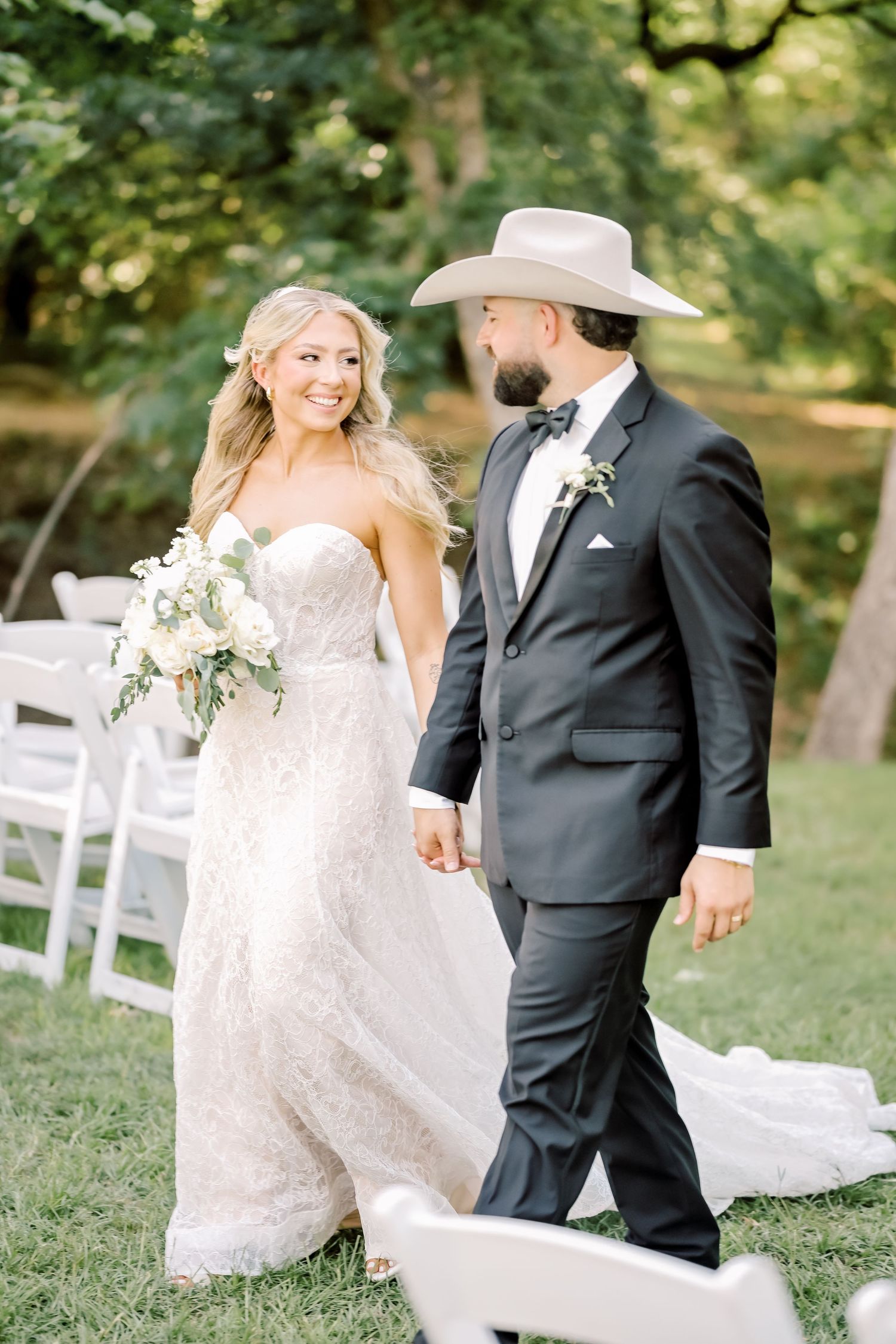 Smiling bride in lace gown and groom in dark suit with cowboy hat walking hand in hand outdoors.