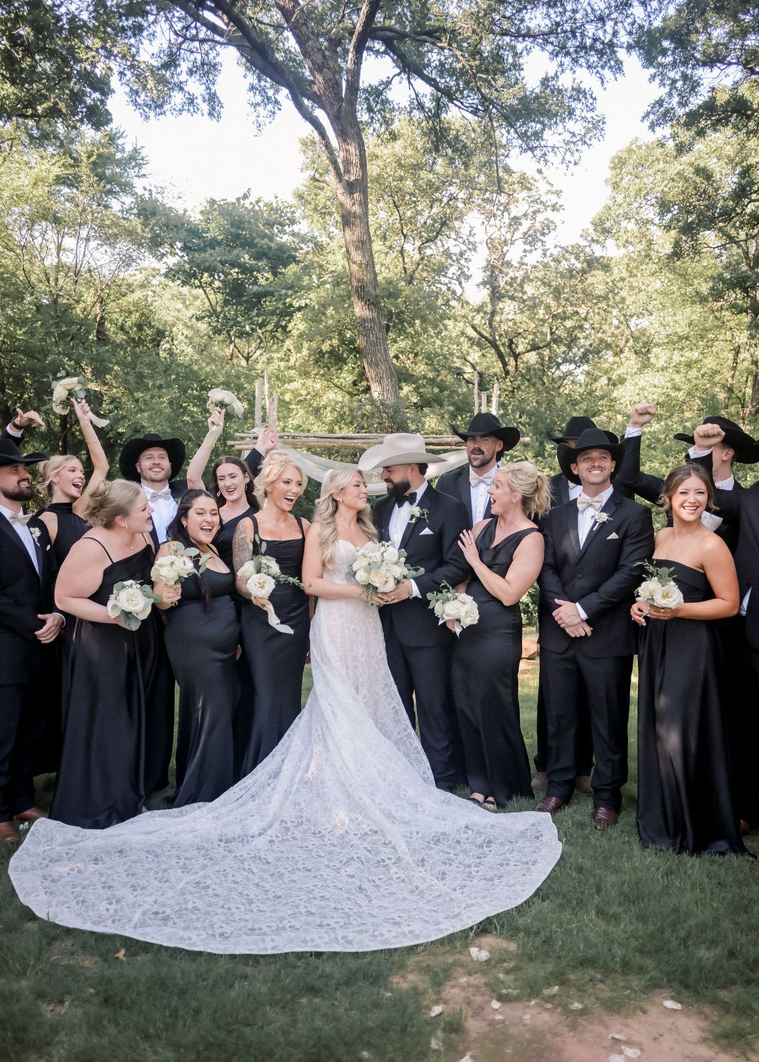 Wedding party in black attire with cowboy hats celebrating with bride in lace gown outdoors.
