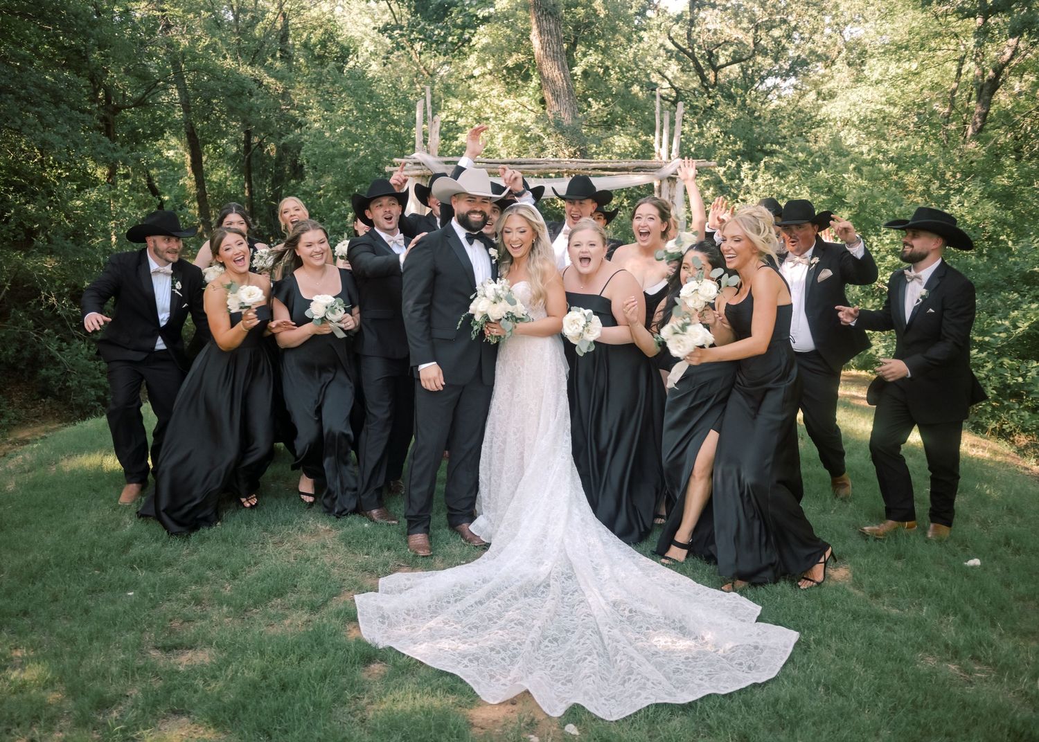 Joyful wedding party in black attire with cowboy hats celebrates with bride and groom outdoors.