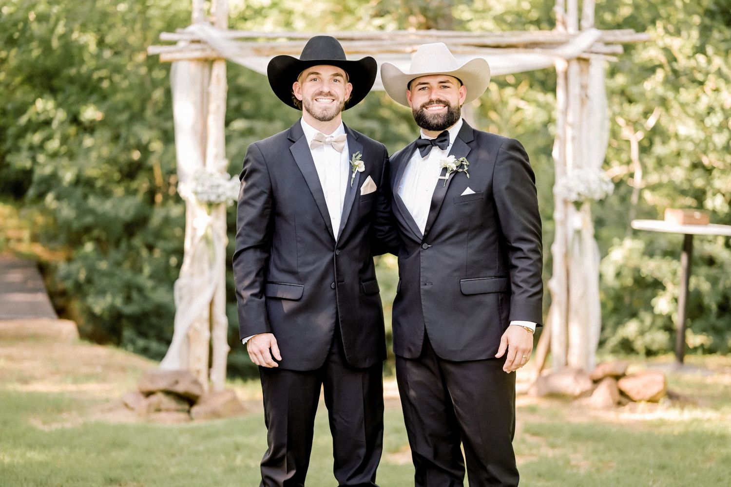 Two men in black suits and cowboy hats pose together at outdoor rustic wedding ceremony altar.