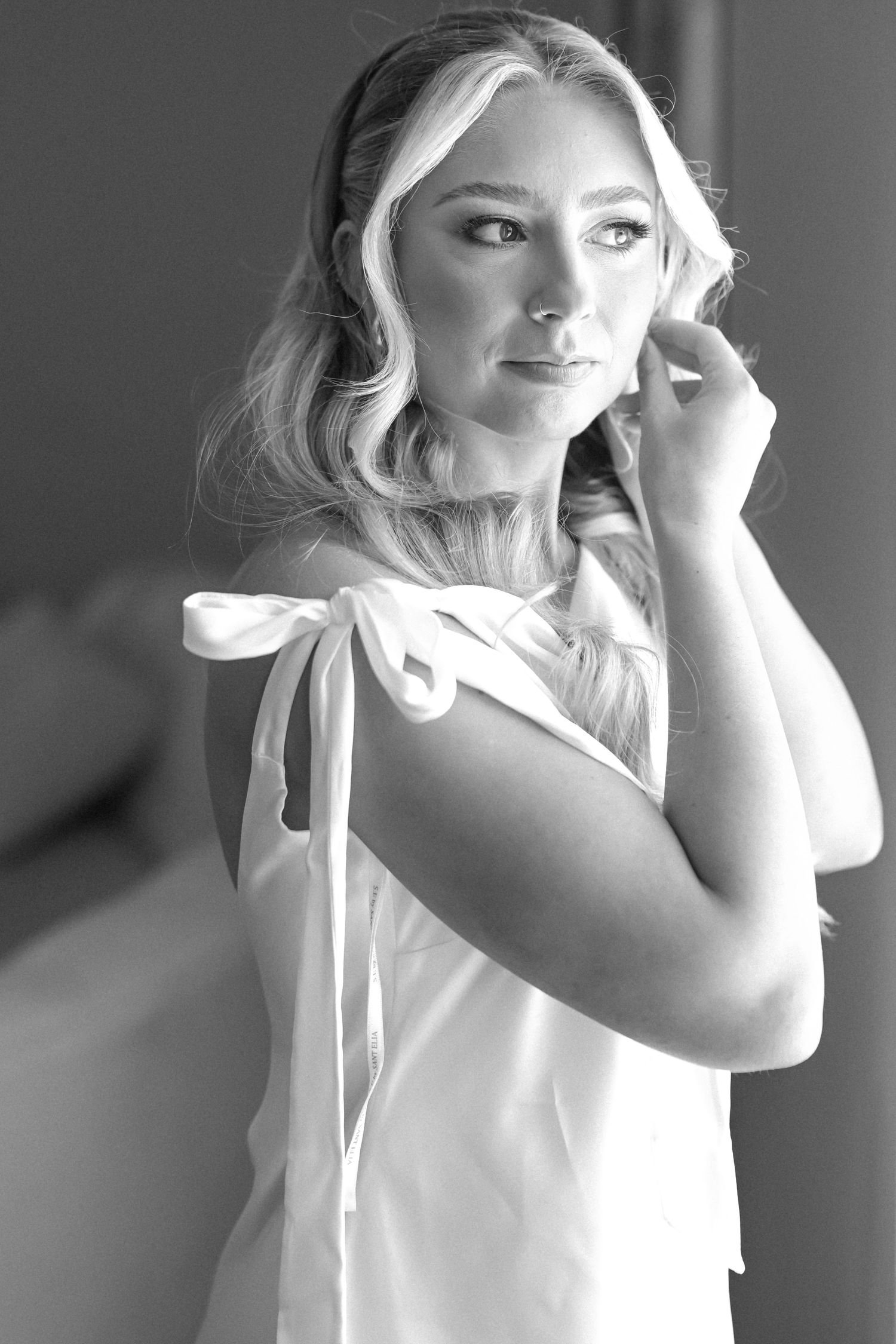 Black and white portrait of a blonde bride in a white dress adjusting her earring.