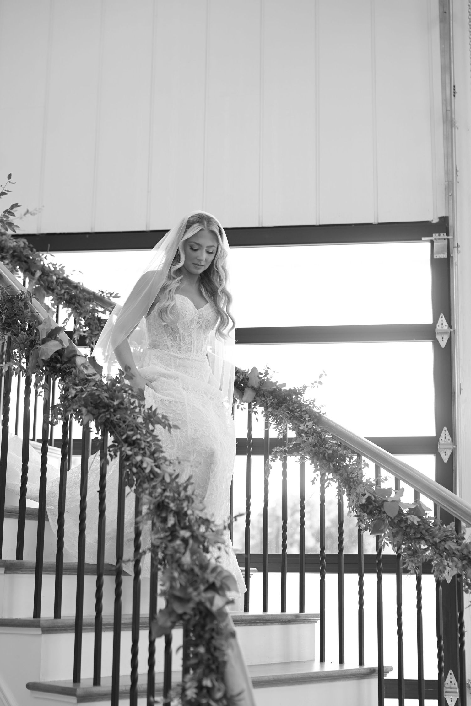 Bride in lace wedding gown and veil descends floral-adorned staircase in black and white wedding photo.